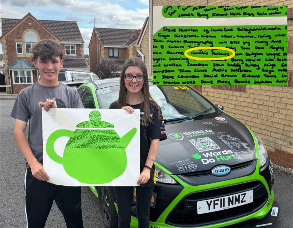 Two people holding a large green teapot cut-out beside the Thomas Potter Racing car