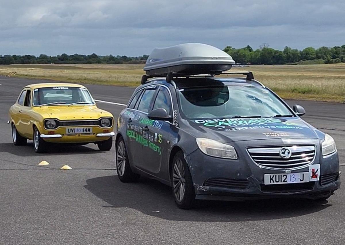 The grey Vauxhall Insignia estate with a homemade cannabis livery parked on grass beside a yellow classic car at Thruxton