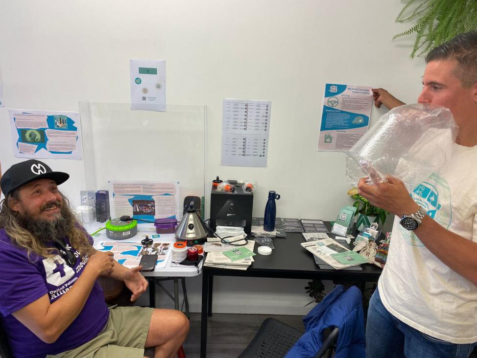 Two people at a PatientsCann UK information stand, one using a vaporiser balloon bag, surrounded by patient information leaflets and posters.